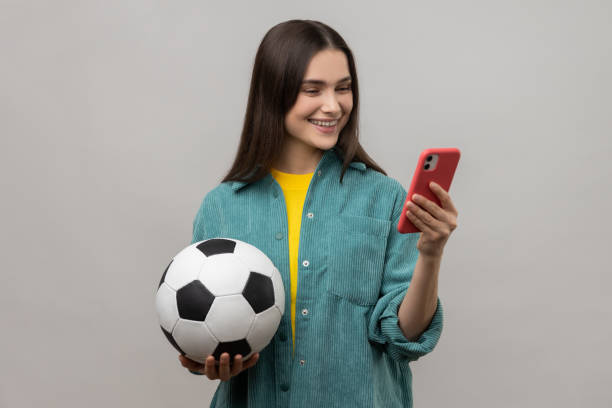 Delighted woman standing holding soccer ball and using smart phone, expressing positive emotions. stock photo