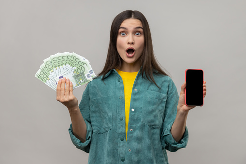 Astonished surprised rich woman holding euro banknotes and phone with blank screen for advertisement or promotion, wearing casual style jacket. Indoor studio shot isolated on gray background.