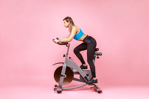 Side view full length of beautiful sportswoman cycling a bike at home, cardio training, exercising legs, wearing sports tights and top. Indoor studio shot isolated on pink background.