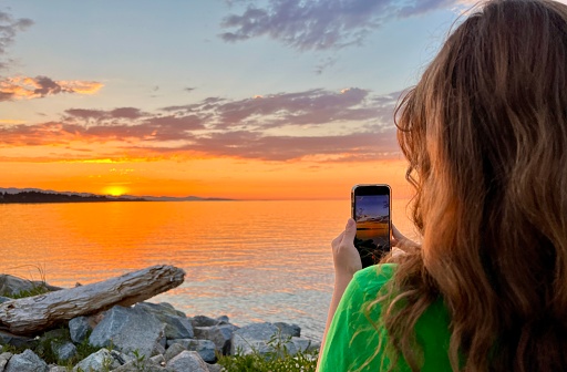 a teenage girl in a Green T-shirt stands with her back to the camera and shoots a beautiful sunset on Pacific Ocean she holds an iPhone 13 phone in her hands blond red hair stones setting sun sea sky