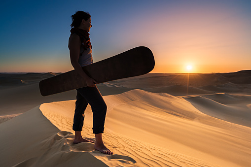 Young female sandboarding in The Sahara Desert during the sunset. She is standing on a sand dune, holding sandboard and watching the sunset over desert.