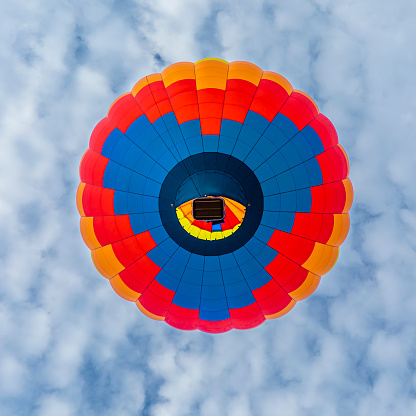 Blue, orange and red hot air balloon directly overhead in a mostly cloudy sky.