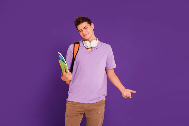 istock Young happy man student in casual clothes posing with backpack and notebooks, isolated on studio violet background. Education concept. 1419155156