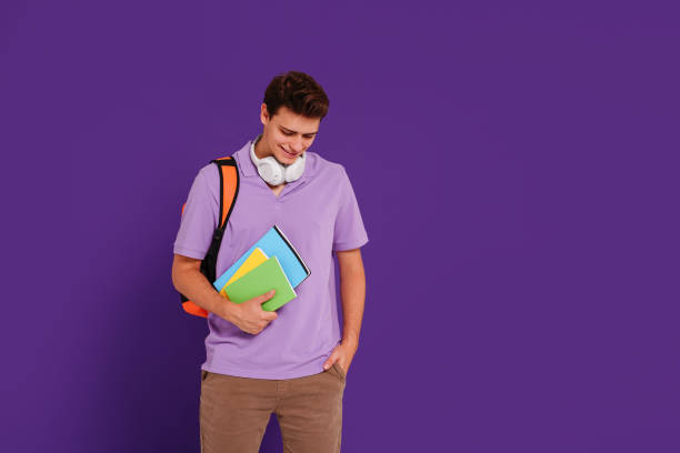 istock Young happy man student in casual clothes posing with backpack and notebooks, isolated on studio violet background. Education concept. 1419155175