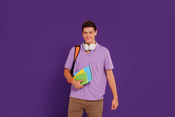 istock Young happy man student in casual clothes posing with backpack and notebooks, isolated on studio violet background. Education concept. 1419155182