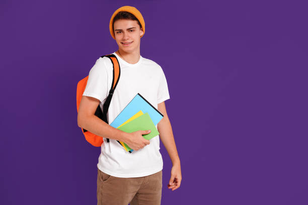 istock Young happy man student in casual clothes and cap, with backpack and notebooks, isolated on studio violet background. Education concept. 1419155192