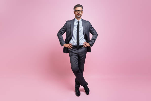 istock Confident mature man in full suit holding hands on hip while standing against pink background 1419155221