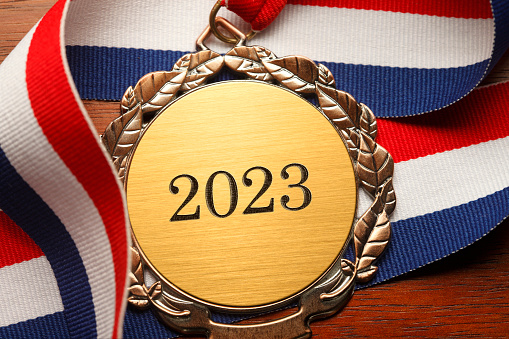 Gold medal engraved with the year 2023 rests on a wood desk framed by red white and blue ribbons.