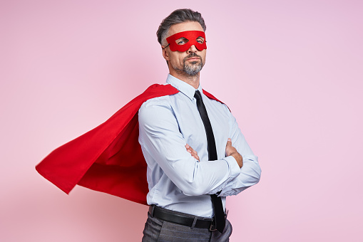 Confident man in shirt and tie wearing superhero cape and keeping arms crossed against pink background