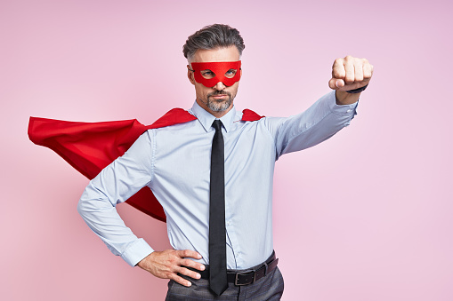 Powerful man in red hero cape posing, showing determination and force to protect people. Smiling strong adult acts as an inspiration or superhero, stands against blue background.