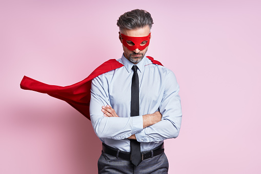 Confident mature man in shirt and tie wearing superhero cape while standing against pink background