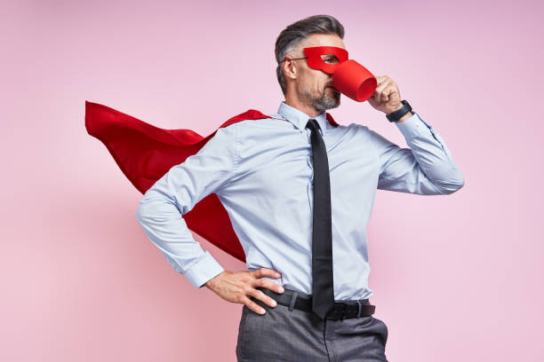 Confident man in shirt and tie wearing superhero cape and drinking hot drink against pink background stock photo