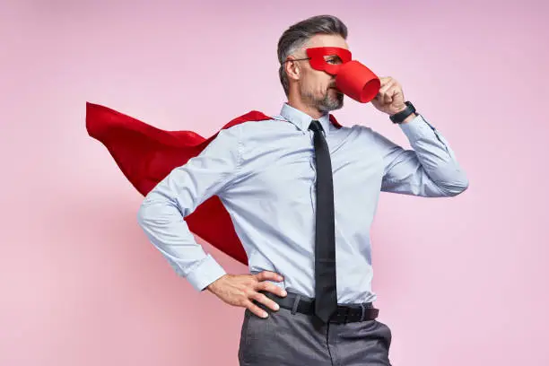 Confident man in shirt and tie wearing superhero cape and drinking hot drink against pink background