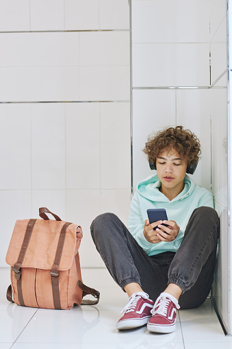 Minimal full length portrait of isolated teen schoolboy sitting alone in corner and using smartphone