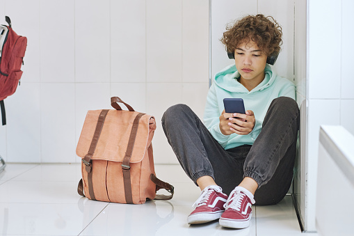 Full length portrait of isolated teen schoolboy sitting alone in corner and using smartphone, copy space