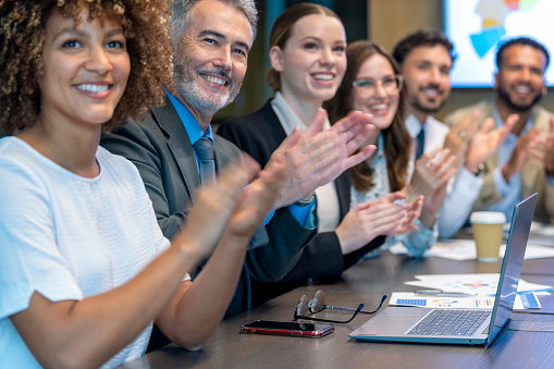 Group of business people applauding a presentation. They are sitting at a conference table. There are several ethnicities present including Caucasian, African and Latino