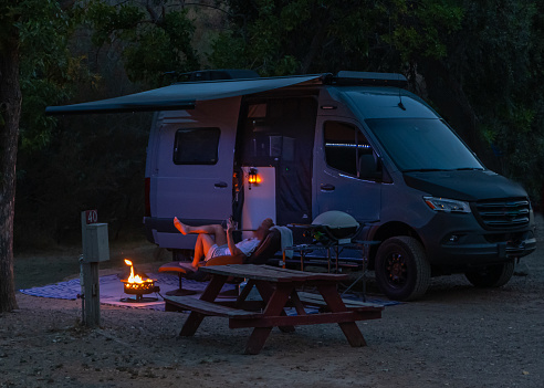 Camper in a camper van is laying next to a fire in the evening at campsite