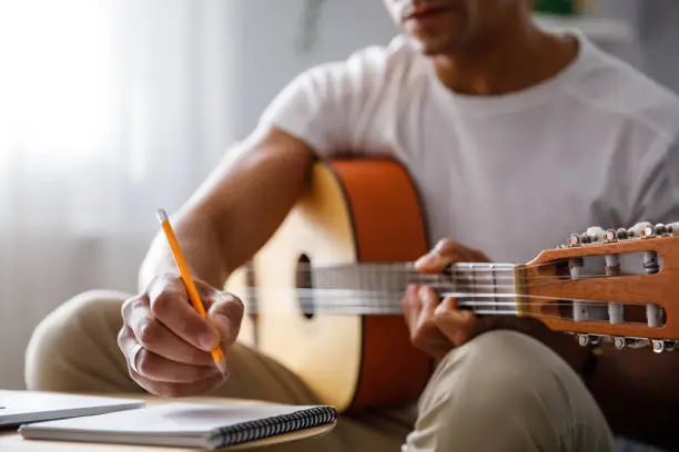 Man composing a song for acoustic guitar, writing chords in a notebook at home