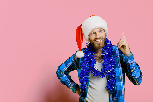 Young adult bearded guy 20s in santa claus hat, new year tinsel makes a gesture with index finger up on a pink isolated background with copy space. Shows gesture idea