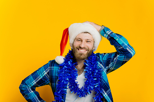 Portrait of a young adult guy 20-30s in tinsel, who straightens the santa hat on his head with his hand and looks at the camera with a smile on a yellow isolated background.