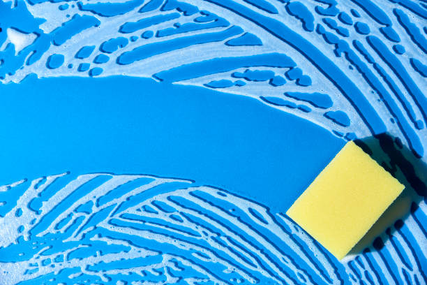 Yellow dishwashing sponge on a blue glass surface covered with soap foam. Top view composition with copy space. Concept of cleanliness.