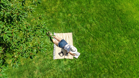 Young woman reading book in park, student girl relaxing outdoors sitting on grass with book and headphones, aerial drone view from above