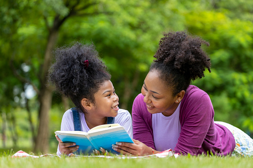 African mother is teaching her young daughter to read while lying down after having a summer picnic in the public park for education and happiness