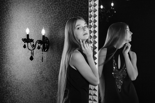Black and white portrait of pretty fashionable teen girl model an elegant dress posing at mirror looking at camera in dark interior of room. Concept of style, fashion and beauty. Copy text space