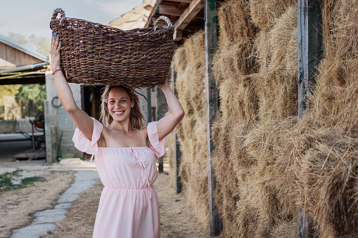 Portrait of happy woman in summer dress working on a farm. She carrying a basket near hay.