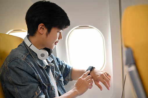 Handsome young Asian male passenger sits at a window seat in economy class, checking a certain time on his wrist watch during flight.