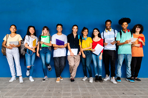 Group of multiracial high school teenage students looking at camera standing on blue background holding binders and wearing backpacks. Back to school concept. Education concept.