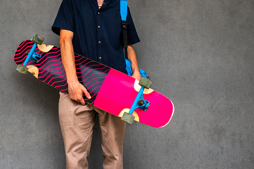 Unrecognisable teen boy holding longboard on dark concrete wall background. Copy space.
