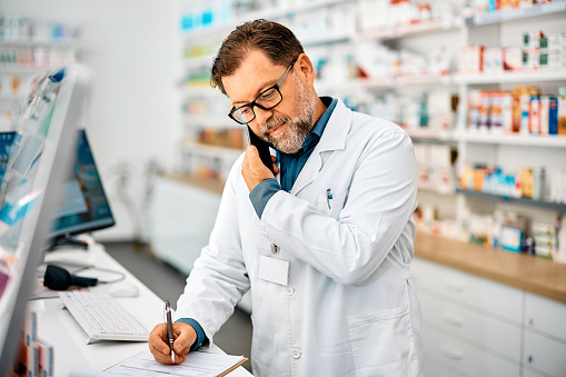 Mature pharmacist communicating on mobile phone and writing notes while working in drugstore.