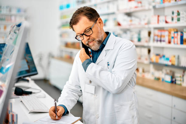 Pharmacist taking notes while talking on the phone in a pharmacy. Mature pharmacist communicating on mobile phone and writing notes while working in drugstore. pharmacist stock pictures, royalty-free photos & images