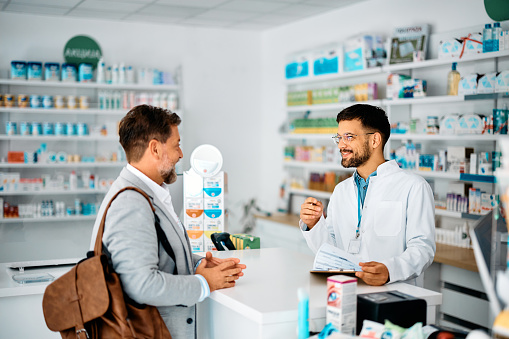 Young happy pharmacist advising a man who is searching for a medicine in drugstore.