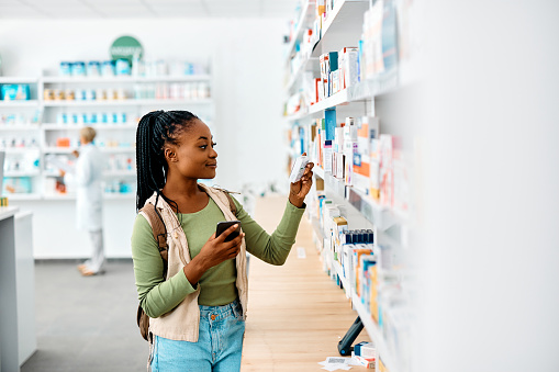 Young African American woman using cell phone while looking for a medicine in drugstore. Copy space.