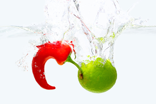 Curled red chilli and whole lime dropped in water with bubbles and splashes isolated on white background.