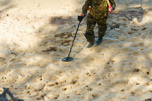 A man with a metal detector walks at the water's edge of a sandy beach. Man with a metal detector on a sea sandy beach searching gold treasure.