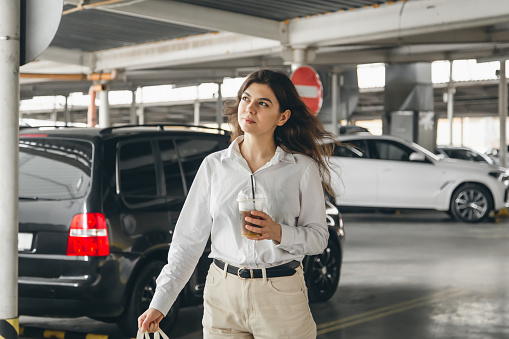 Attractive business young woman in a white shirt in a car park with a cup of coffee.