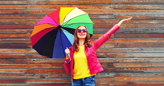 Autumn portrait of happy smiling young woman holding colorful umbrella wearing red jacket, heart shaped sunglasses on city street on wooden wall background