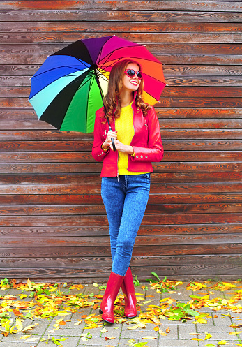 Autumn portrait of happy smiling young woman holding colorful umbrella wearing red jacket, rubber boots, heart shaped sunglasses on city street on wooden wall with yellow leaves background