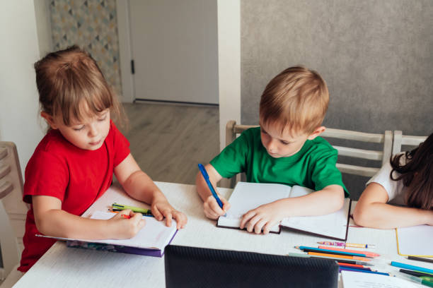 Children sit at a table and write in notebooks and use a laptop