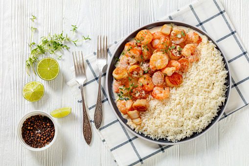 shrimps and scallops in spicy coconut cream sauce with long grain rice on plate on white wooden table with forks and lime,  horizontal view from above, flat lay