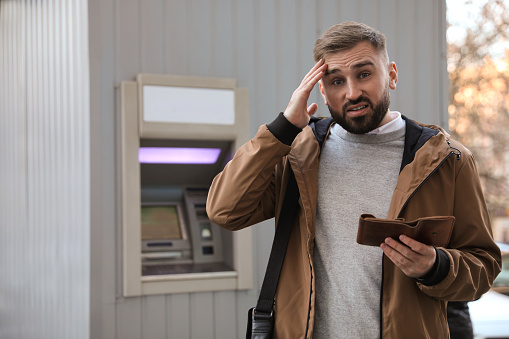Unhappy young man with wallet near cash machine outdoors