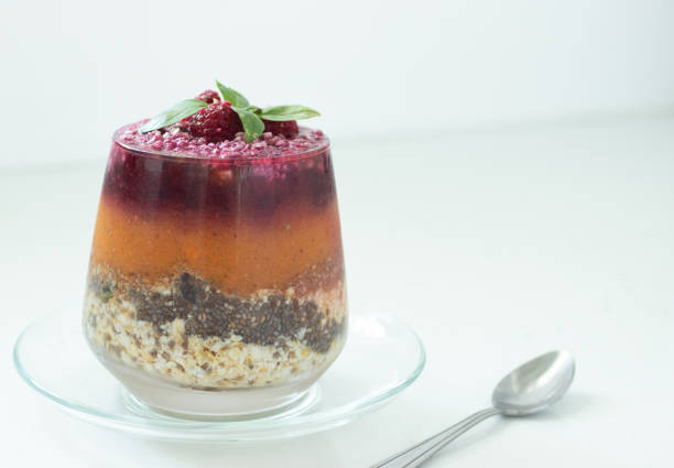 Layered plant-based chia pudding made with fresh fruit, veggies, oats, chia seeds, and nuts in a glass with a spoon on white. A closeup.