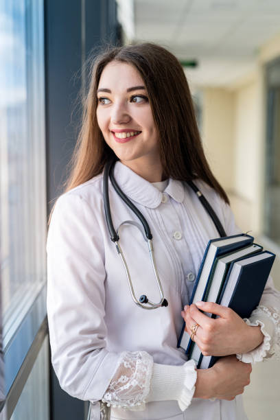 istock smiling young female doctor in uniform with stethoscope holding book while standing 1419279889