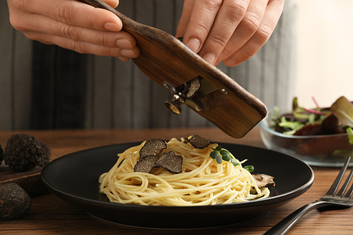 Woman slicing truffle onto spaghetti at wooden table, closeup
