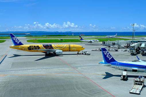 Naha, Japan - August 4, 2022: An All Nippon Airways 777 with C-3PO Star Wars livery leaves a jet bridge at Naha Airport, Okinawa behind a parked ANA A321neo in the airline's regular blue and white livery. Despite Japan's seventh and largest surge in COVID-19 cases, this year's summer travel season for domestic travel has been the busiest since the pandemic began.