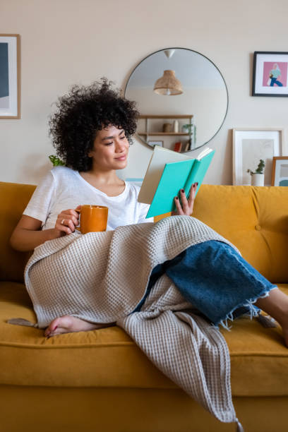 mulher afro-americana relaxada e feliz na sala de estar de casa sentada no sofá, lendo um livro e bebendo chá - resting relaxation book break - fotografias e filmes do acervo