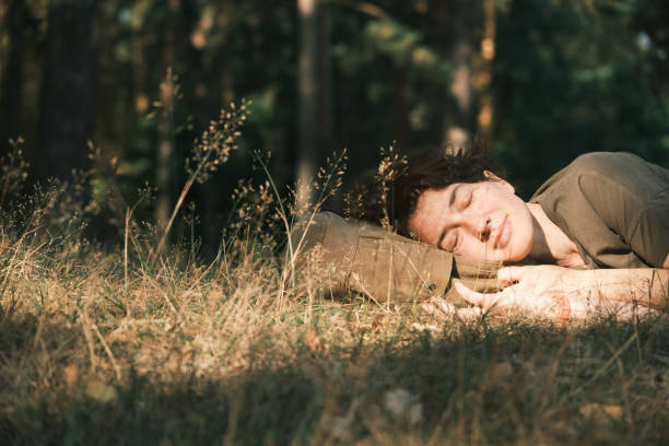 A man with a beard lies back on green grass, hands behind his head, eyes closed. He is wearing a bright yellow shirt and seems to be enjoying a calm afternoon outdoors.
